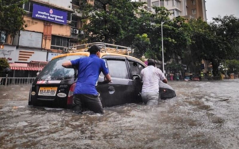 Natural disaster mass flooding. 3-4 men push car up village street in waist high raging flood water.