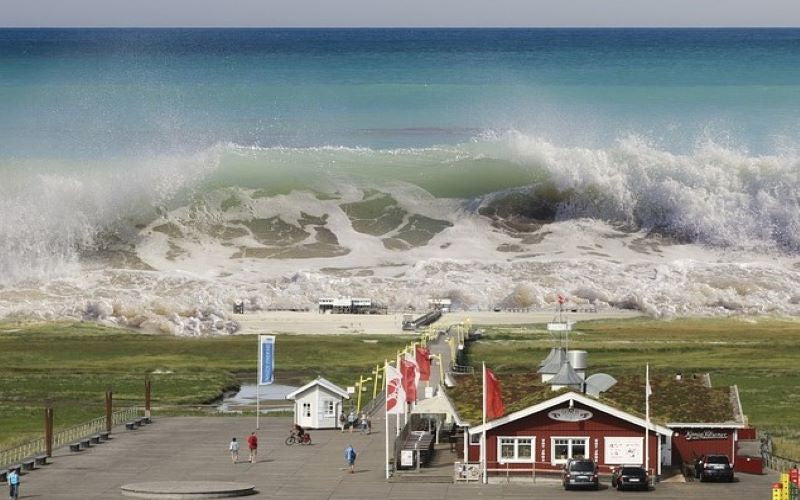 Aerial photo of inbound tsunami seaquake wave across grass field, red building, and seaside beach huts. St Peter-Ording, Germany 2016